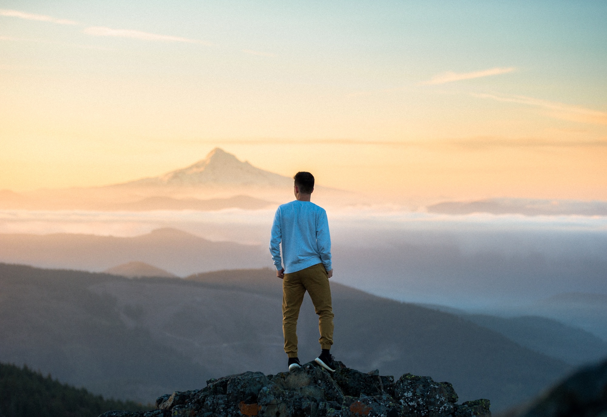 Man atop rocky peak looking away at sunset behind distant mountain.
